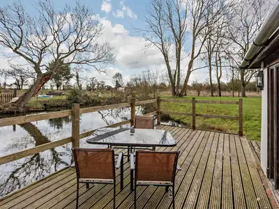 a table and chairs on a wooden deck next to a river at Pond View Cottage - Uk48230 in Tibenham