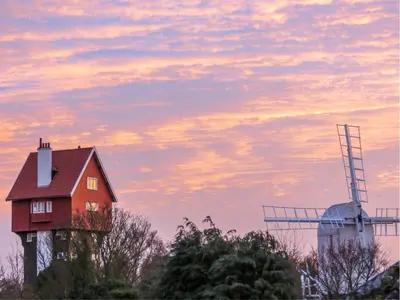 a red house and a windmill with a cloudy sky at 2 Bell Lodge, Thorpeness in Thorpeness